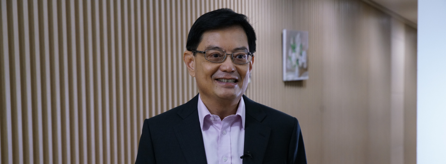 Heng Swee Keat in a suit and glasses stands in front of a textured wall with artwork.